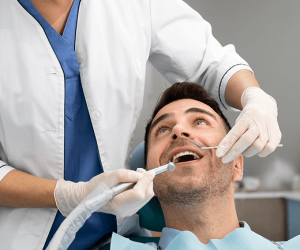 Mann sitzt auf einem Zahnarztstuhl und lächelt, während eine Zahnärztin mit Mundschutz und Handschuhen seine Zähne untersucht.